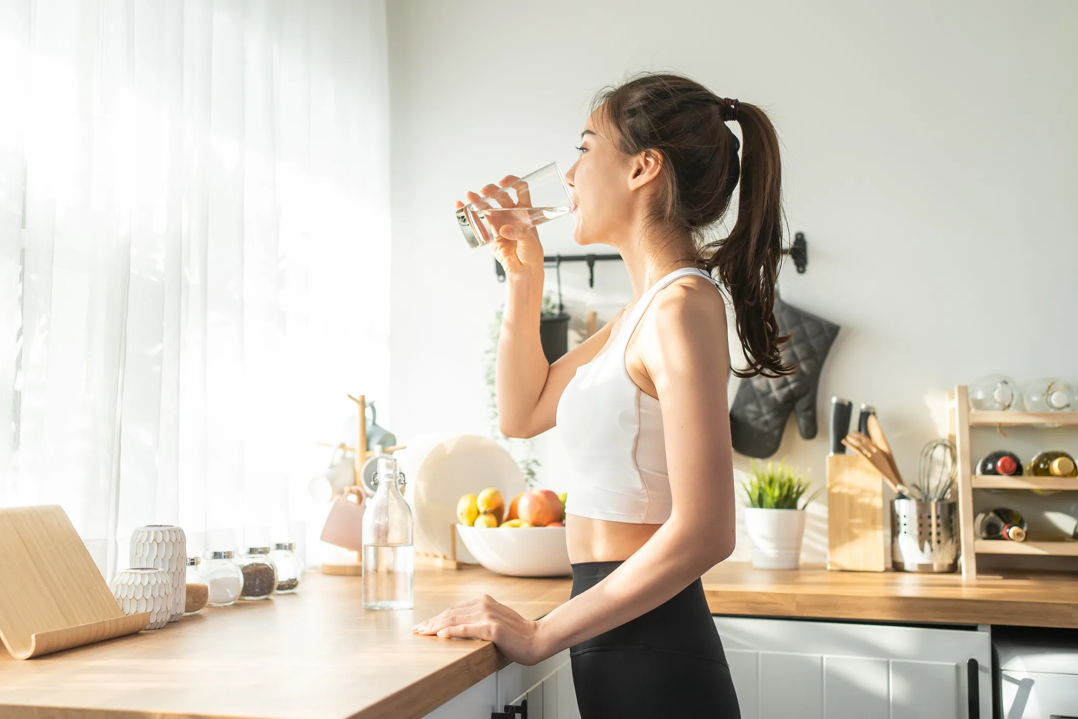 Asian beautiful woman in sportswear drinking hydrogen water after exercise at home. Young thirsty active sport girl takes a sips of clean mineral natural in cup after workout for health care in kitchen in house.