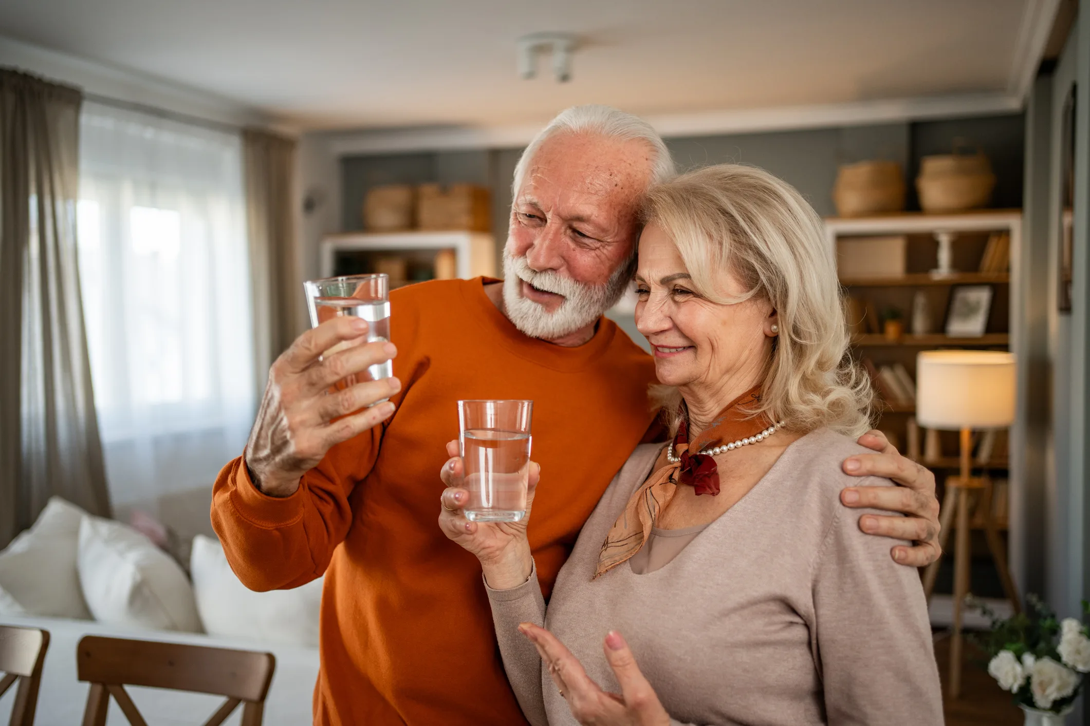 An elderly couple is standing in the living room drinking water and taking care of their health. Their smiles reflect the satisfaction of a moment of health and togetherness. The scene conveys a sense of self-care and the simple joy of sharing everyday moments of love and care late in life.